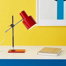 Load image into Gallery viewer, A modern red and black adjustable table lamp with a rectangular orange base sits on a white desk against a bright yellow wall, next to a closed beige notebook.
