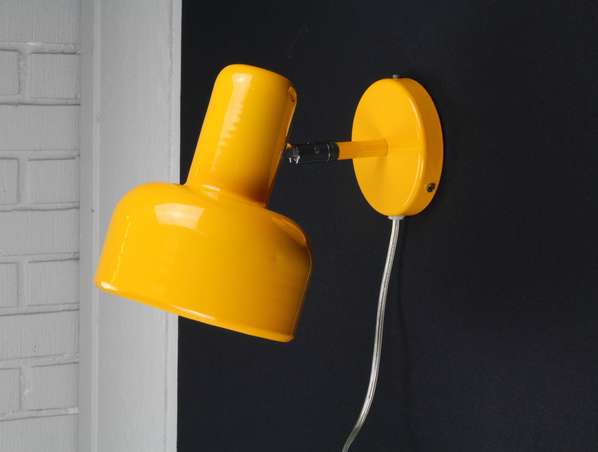 Yellow wall lamp attached to a black wall with a gray brick wall in the background
