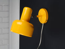 Load image into Gallery viewer, Yellow wall lamp attached to a black wall with a gray brick wall in the background
